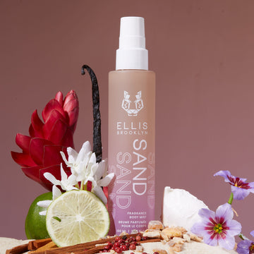 Sand Hair and Body Fragrance Mist 100ml in sand with white, purple, and red flowers, bergamot, vanilla beans, sandal wood, pink pepper, and coconut, against purple and brown gradient background.