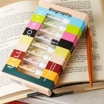 Biography Discovery Set box showing Salt 1.5ml Eau De Parfum Vial, Sun Fruit 1.5ml Eau De Parfum Vial, Florist 1.5ml Eau De Parfum Vial, Myth 1.5ml Eau De Parfum Vial, Sweet 1.5ml Eau De Parfum Vial, Vanilla Milk 1.5ml Eau De Parfum Vial, Super Amber 1.5ml Eau De Parfum Vial, Bee 1.5ml Eau De Parfum Vial, and Apres 1.5ml Eau De Parfum Vial, resting on opened book, stacked on three closed books, next to orange pen.