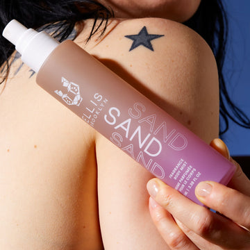Model with black hair and tattoos on shoulder holding Sand Hair and Body Fragrance Mist 100ml without cap on back of shoulder, on blue background.