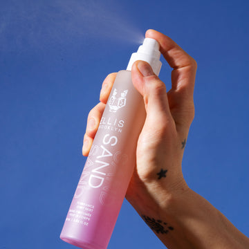 Model with hand and wrist tattoos spraying Sand Hair and Body Fragrance Mist 100ml, on blue background.