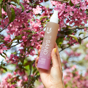 Model with yellow nail polish wearing gold ring holding Sand Hair and Body Fragrance Mist 100ml in front of tree branches with pink flowers, on blue background.