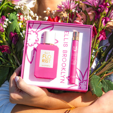 Model with blonde hair and nude nail polish, wearing white long sleeve shirt and gold bracelets holding Florist Eau De Parfum Gift Set Box showing Florist 50ml Eau De Parfum and Florist 10ml Eau De Parfum Travel Spray and bouquets of red, pink, and white flowers in both arms against chest.