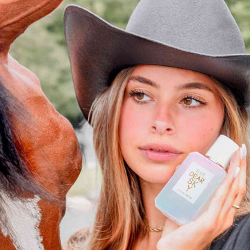 Model with brown hair and white nail polish, wearing black shirt, brown cowboy hat, gold necklace, and silver ring holds Dear Sky 50ml Eau De Parfum next to face, while standing next to brown and white horse with black hair, with trees and road in the background.