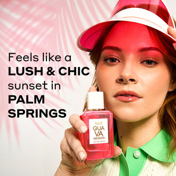 Model with red hair, nude nail polish, and gold nose ring wearing green and white collared polo shirt and pink and white visor holding Guava Granita 50ml Eau De Parfum against white background with pink palm leaf shadow and black text that reads "Feels like a lush & chic sunset in Palm Springs".