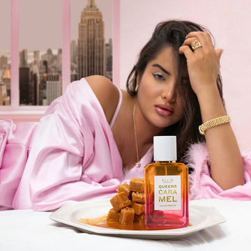 Queens Caramel 50ml Eau De Parfum on white plate with blocks of caramel and melted caramel, on white bed, in front of model with black hair wearing gold ring, gold earrings, gold bracelet, gold necklace, and pink satin robe with feathers at cuffs laying on side, on background of pink wall and pink window with view of buildings in New York City.