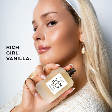 Model with blond hair and nude nail polish wearing white headband, pearl earring, gold rings, and cream boucle shirt holding Vanilla Salt 50ml Eau De Parfum without cap near shoulder on white background with black text that reads "Rich Girl Vanilla."