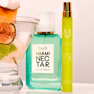 Miami Nectar 50ml Eau De Parfum and Sun Fruit 10ml Eau De Parfum Travel Spray next to glass filled with sparkling water, with apple, fig, pink pineapple, and coconut on skewer, on white table, on white background.