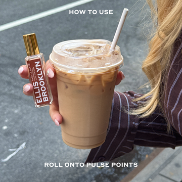 Model with nude nail polish and blonde hair wearing a brown and white stripped long sleeve button-down shirt holding and iced coffee and Vanilla Santo Fragrance Oil 15ml in one hand on city street between white text that reads "How To Use Roll Onto Pulse Points".