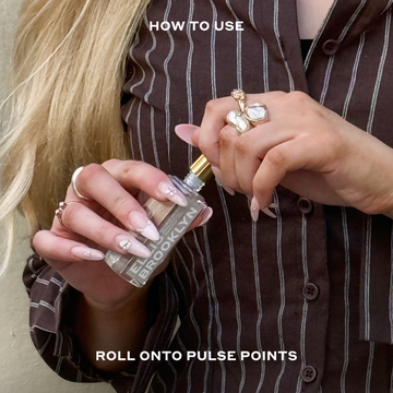 Model with nude nail polish and blonde hair wearing a brown and white stripped long sleeve button-down shirt and assorted Peral rings removing cap of Blond Musk Fragrance Oil 15ml against beige background between white text that reads "How To Use Roll Onto Pulse Points".