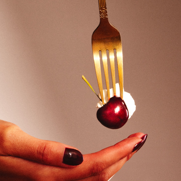 Model with red nail polish holding hand under gold fork with cherry and whipped cream, on tan background.