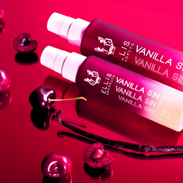 Vanilla Sin Hair And Body Fragrance Mist 100ml behind Vanilla Sin Hair And Body Fragrance Mist 50ml on red mirror next to cherries and vanilla bean.