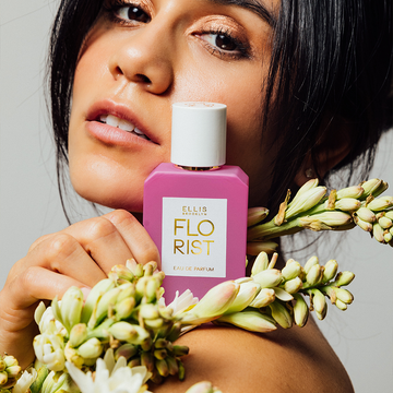 Model with black hair pulled back wearing diamond earring holding Florist 50ml Eau De Parfum and flowers on shoulder against light grey background.