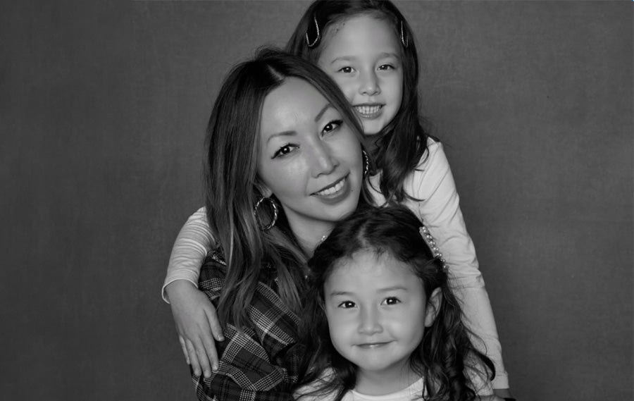 Bee Shapiro wearing hoop earrings, necklace, and plaid shirt holding both her daughters wearing white long sleeve shirts and clips in hair, on grey background.