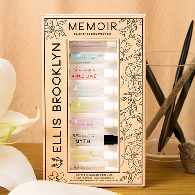Memoir Fragrance Discovery Set box with beige and black floral design showing Dear Sky Eau De Parfum 1.5ml Vial, Miami Nectar Eau De Parfum 1.5ml Vial, Apple Love Eau De Parfum 1.5ml Vial, Salt Eau De Parfum 1.5ml Vial, Sun Fruit Eau De Parfum 1.5ml Vial, Florist Eau De Parfum 1.5ml Vial, Myth Eau De Parfum 1.5ml Vial, Sweet Eau De Parfum 1.5ml Vial, and Vanilla Milk Eau De Parfum 1.5ml Vial, on wooden table leaning against glass of black and brown pens, next to white flowers, against beige background.