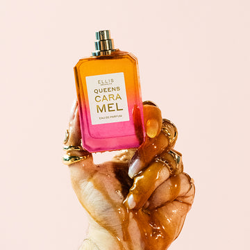 Model with nude nail polish wearing gold rings with hand covered in melted caramel holding Queens Caramel 50ml Eau De Parfum 50ml without cap, on light pink background.