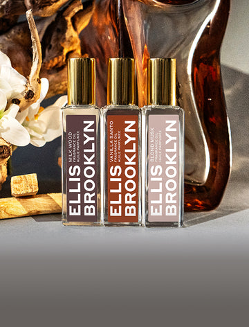 Milk Wood Fragrance Oil 15ml, Vanilla Santo Fragrance Oil 15ml, and Blond Musk Fragrance Oil 15ml in front of sandal wood, block of amber, and piece of wood with white flowers, on grey background.