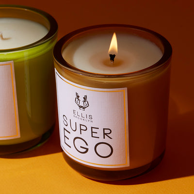 Superego 6.5 oz Terrific Scented Candle with wick lit next to Fable 6.5 oz Terrific Scented Candle, on orange background.
