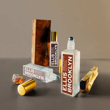 Vanilla Santo Fragrance Oil 15ml with cap removed next to palo santo, Blond Musk Fragrance Oil 15ml with cap removed, blond amber, Milk Wood Fragrance Oil 15ml, and piece of wood against brown backdrop.