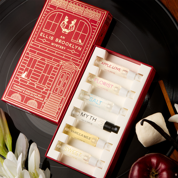 Red An Ellis Brooklyn Mystery Fragrance Discovery Set box opened to show inside and Apple Love 1.5ml Eau De Parfum Vial, Florist 1.5ml Eau De Parfum Vial, Salt 1.5ml Eau De Parfum Vial, Myth 1.5ml Eau De Parfum Vial, Vanilla Milk 1.5ml Eau De Parfum Vial and Sweet 1.5ml Eau De Parfum Vial on black vinyl record with vanilla beans, marshmallows, apple, sandalwood and white flowers, on wooden table.
