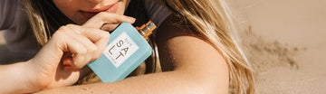 Model with blonde hair wearing white shirt laying down on sand holding Salt 50ml Eau De Parfum without cap with one hand on arm.