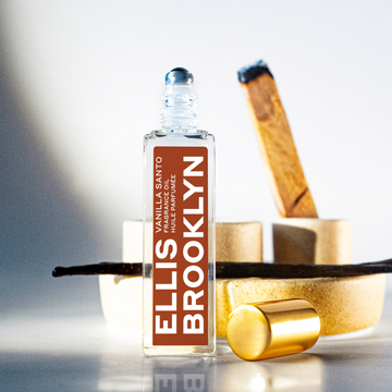 Vanilla Santo Fragrance Oil 15ml with cap removed next to vanilla bean, palo santo, and wooden props on glass table against grey and white background.