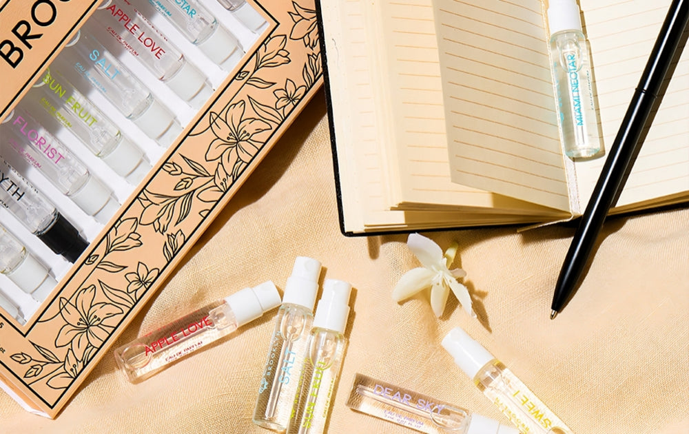 Memoir Fragrance Discovery Set box with beige and black floral design showing Dear Sky Eau De Parfum 1.5ml Vial, Miami Nectar Eau De Parfum 1.5ml Vial, Apple Love Eau De Parfum 1.5ml Vial, Salt Eau De Parfum 1.5ml Vial, Sun Fruit Eau De Parfum 1.5ml Vial, Florist Eau De Parfum 1.5ml Vial, Myth Eau De Parfum 1.5ml Vial, Sweet Eau De Parfum 1.5ml Vial, and Vanilla Milk Eau De Parfum 1.5ml Vial, on tan fabric next to white flowers, and notebook with pen.