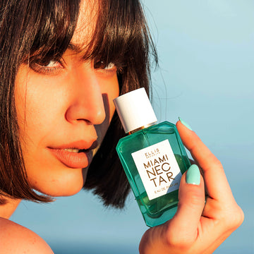 Model with short black hair and teal nail polish holding Miami Nectar 50ml Eau De Parfum near face against blue background.