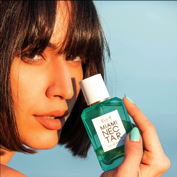 Model with brown hair holding blue Miami Nectar bottle against a blue background.