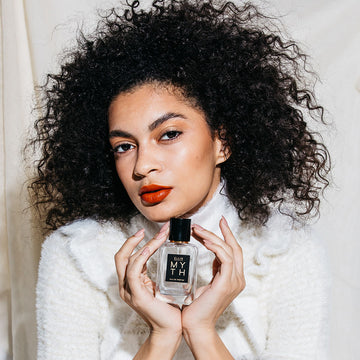 Model with black curly hair, nude nail polish, red lipstick, wearing white sweater against white fabric background holding Myth 50ml Eau De Parfum with both hands near chin.