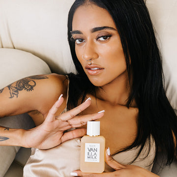 Model with black hair, white nail polish, facial piercings, and dragon tattoo on arm wears beige satin tank top while laying down on off-white couch and holding Vanilla Milk 50ml Eau De Parfum with both hands.