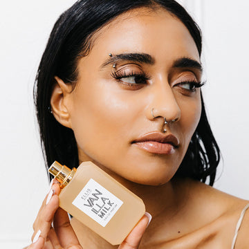 Model with black hair, facial piercings, and white nail polish wearing white tank top holding Vanilla Milk 50ml Eau De Parfum with cap removed near jaw against white background.