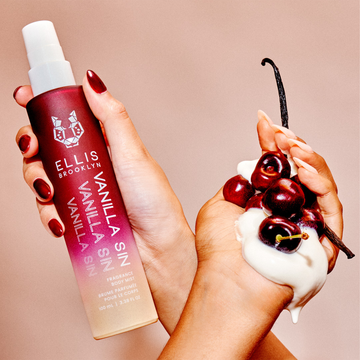 Model with red nail polish holding Vanilla Sin Hair And Body Fragrance Mist 100ml behind model with nude nail polish holding whipped cream, cherries, and vanilla beans, on tan background.