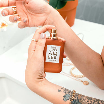 Model with nude nail polish and tattoo on arm wears gold rings and bracelet, holding Super Amber 50ml Eau De Parfum with cap removed and finger on sprayer in front of a white vanity with gold jewelry and a potted flowering plant.