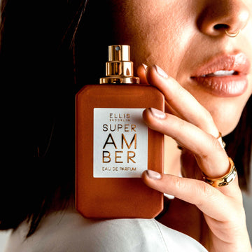 Model with brown hair, nude nail polish and nose ring wearing a white shirt and gold rings holding Super Amber 50ml Eau De Parfum with cap removed on shoulder.