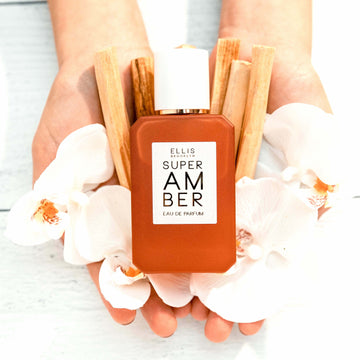 Hands holding Super Amber 50ml Eau De Parfum, white flowers, and pieces on wood against white wooden table.