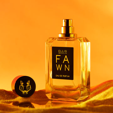 Fawn 50ml Eau De Parfum with cap removed on mirrored surface with sand, on orange background.
