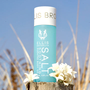 Salt Soothing Scented Body Oil 95ml next to white flowers on piece of wood with beach dunes and sky in background.