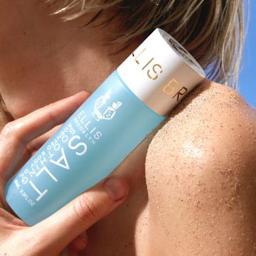 Model with short blonde hair holding Salt Soothing Scented Body Oil 95ml against sandy shoulder with blue background.