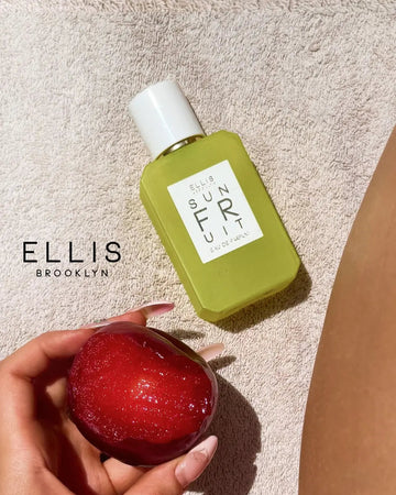 Sun Fruit 50ml Eau De Parfum on creme towel in between Ellis Brooklyn logo, and leg of model, above hand with French Manicure holding an apple.