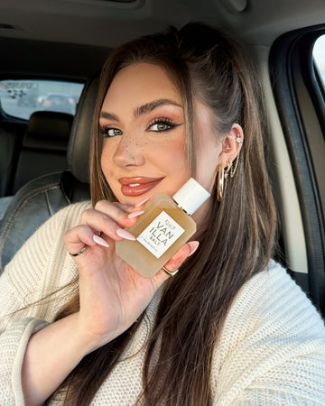 Model with brown hair in a ponytail wearing a white sweater and gold earrings and rings sits in the front seat of a car and faces camera while holding Vanilla Salt Eau de Parfum 50ml