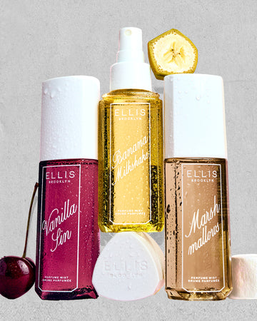 VANILLA SIN Perfume Mist 240ml next to a cherry, BANANA Milkshake Perfume Mist 240ml sitting on top of it's cap, MARSHMALLOWS Perfume Mist 240ml with a slice on banana on top of it and a marshmallow next to it, all covered in water droplets on a gray, rough background 