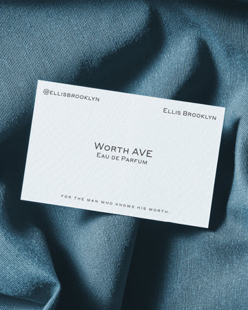 White business card with black text that reads "@ellisbrooklyn Ellis Brooklyn Worth Avenue Eau De Parfum For The Man Who Knows His Worth." on blue fabric.