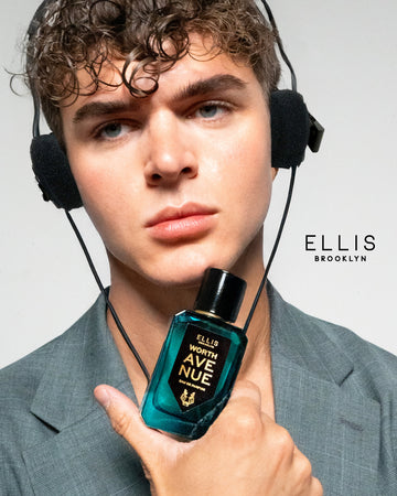Model with brown hair wearing gray jacket and headphones holding Worth Avenue 50ml Eau De Parfum to chin, on white background with black Ellis Brooklyn logo.