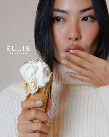Model with brown hair and nude nails wearing white sweater holding vanilla ice cream cone with gold and diamond jewelry on top, while holding other hand to mouth, on gray background with white Ellis Brooklyn logo.