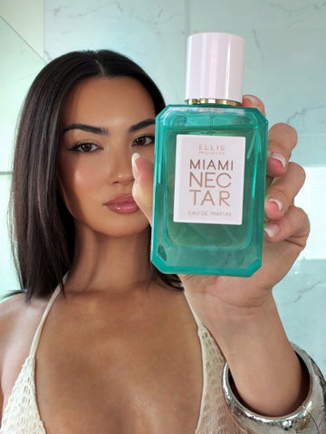 Woman with brown hair wearing a white crocheted bikini top and a silver bracelet holds MIAMI NECTAR Eau de Parfum 50ml in front of her with white marble in the background