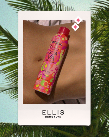 White polaroid with black text that reads "Ellis Brooklyn" of Amika x Ellis Brooklyn GUAVA GRANITA Perk Up Dry Shampoo laying on stomach on top of a white and brown striped towel with overlaid pink and white sparkles on a background of a blue sky and green palm leaves
