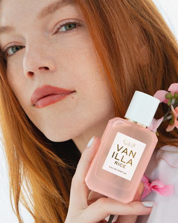 Woman with red hair and a pale pink manicure wearing a pink shirt with bows holds VANILLA RICE Eau de Parfum 50ml and sakura blossoms on white background