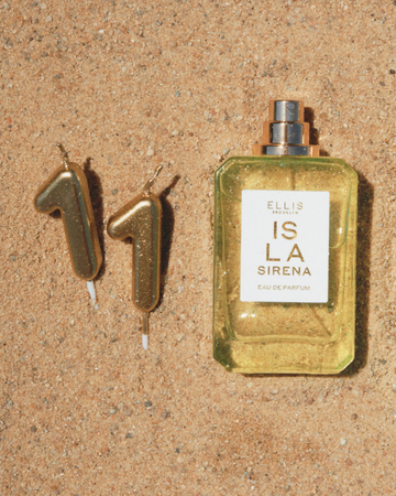 ISLA SIRENA Eau de Parfum 50ml without cap lays in tan sand next to gold candles shaped like the number eleven