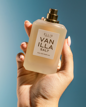 Hand with light pink manicure holds VANILLA SALT Eau de Parfum 50ml without cap on blue background
