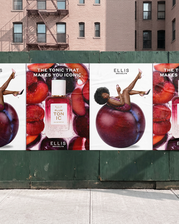 A large outdoor billboard advertisement for Ellis Brooklyn with 'The Tonic That Makes You Iconic' displayed on a green construction wall in front of a brick building. The ad features PLUM TONIC Eau de Parfum 50ml surrounded by sliced red plums and a woman with brown hair wearing a purple sequin dress sitting on a whole plum and wearing pink high heels.